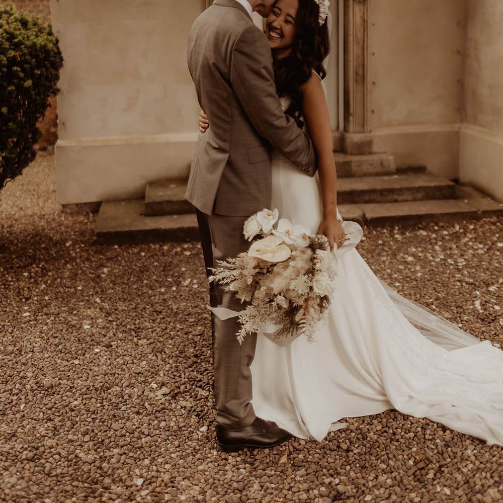 Bride with neutral white wedding bouquet and bridal headband hugs the groom in neutral wedding suit 