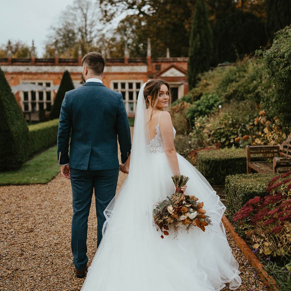 Bride wearing a layered tulle wedding skirt and sequinned top wedding dress looks back at the camera walking with the groom in a blue suit 