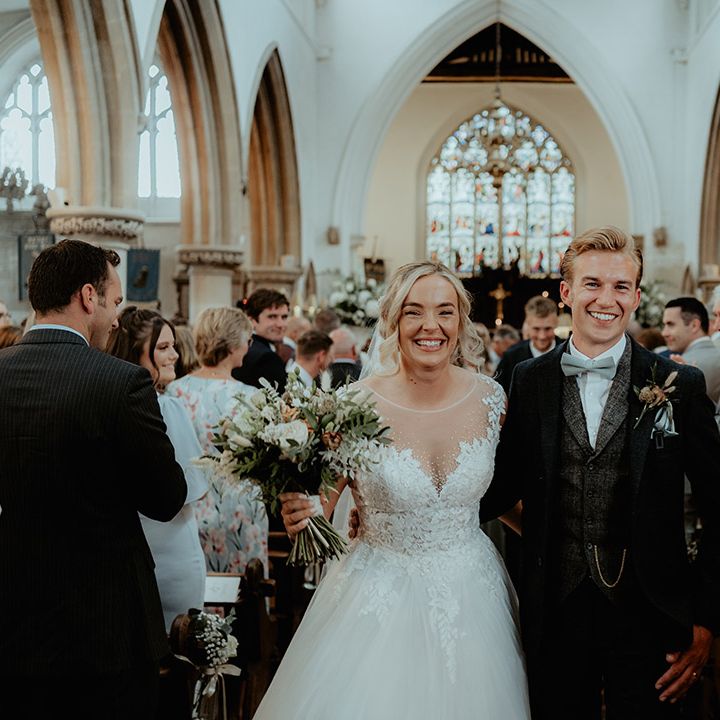 Groom wearing a black suit with a grey tweed waistcoat and sage green bow tie walks back down the aisle with the bride in an illusion princess wedding dress