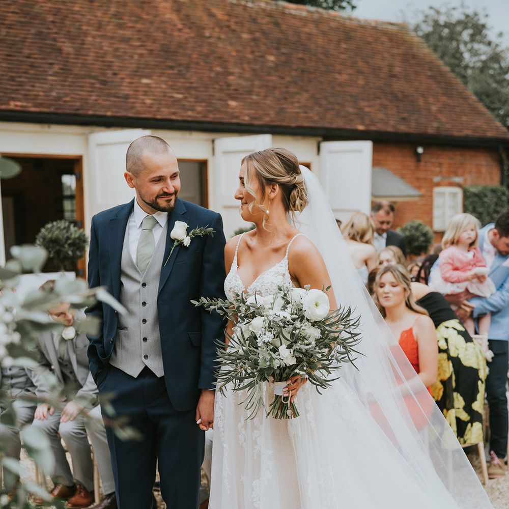 Bride in a floral lace wedding dress standing holding hands at the altar with the groom in a dark blue suit, grey waistcoat and green tie with a white rose buttonhole