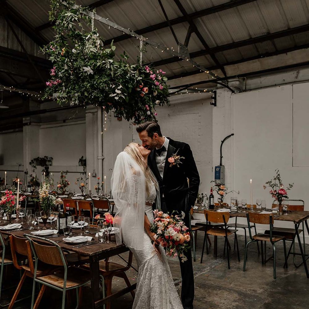 Bride in boho two piece lace bridal outfit with large mixed colourful bouquet and groom in black tux leaning on table at Hackney Studios