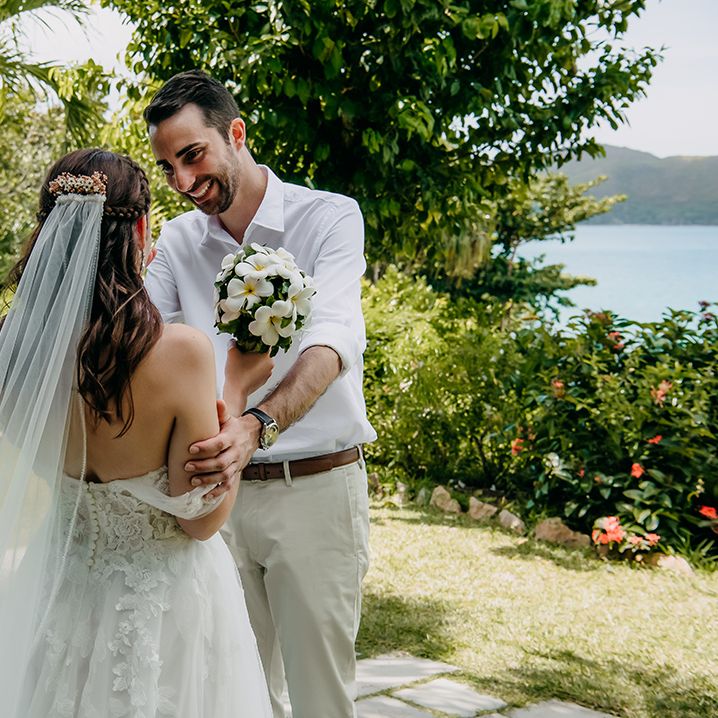 Groom in crisp white shirt, light chinos and tan leather belt embracing bride in off the shoulder lace wedding dress with lace floral details and cathedral length veil