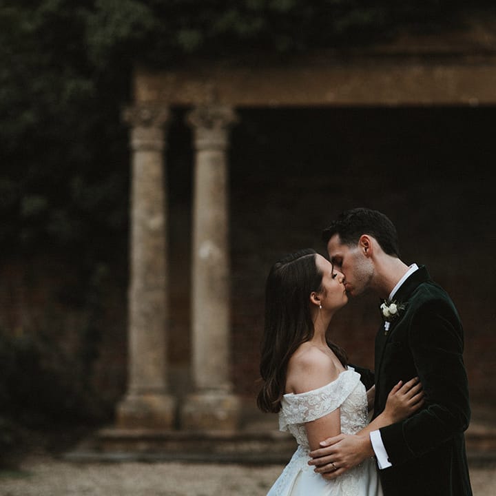 Bride in off the shoulder wedding dress kisses the groom in a dark green tuxedo 