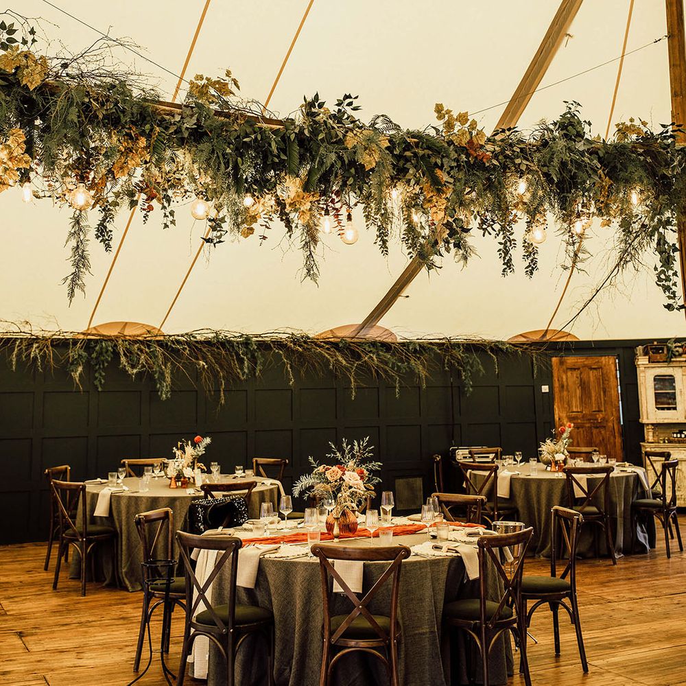 Hanging green foliage installation above round tables with green tablecloths and orange table runners 