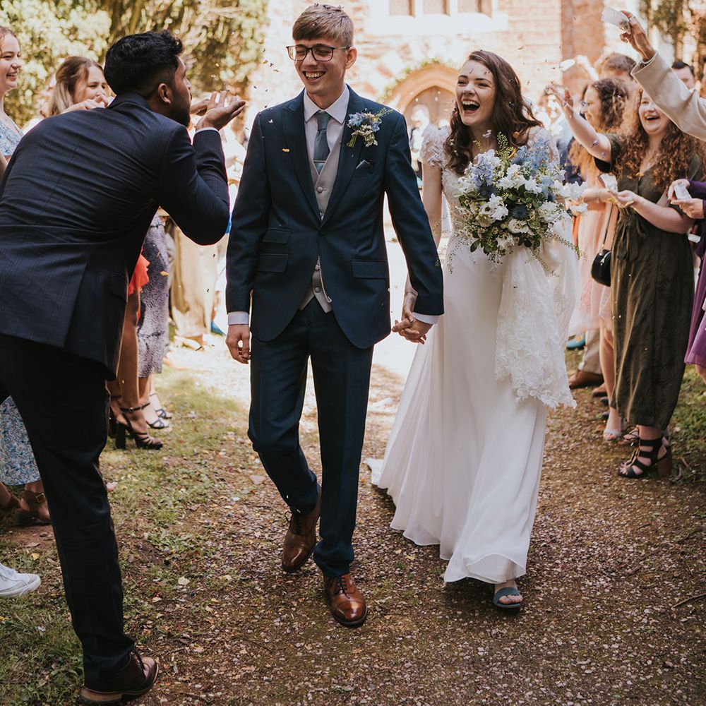 Groom in navy wedding suit with light blue tie and gray waistcoat walking with bride in lace wedding dress for confetti moment  