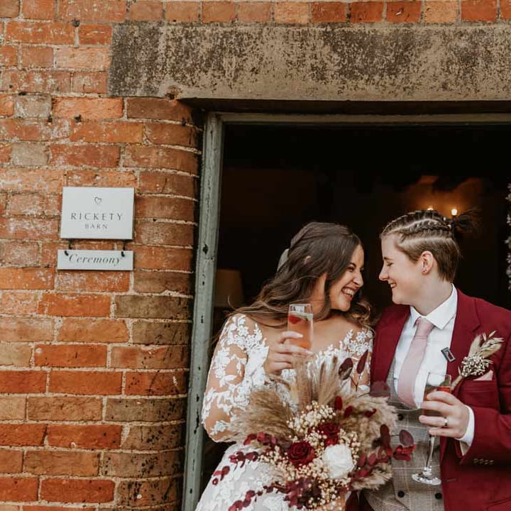 Bride in maroon bridal suit with grey waistcoat, dusky pink tie and autumnal dried flower boutonniere sipping champagne and laughing with bride in long sleeve lace illusion sleeve wedding dress with overlay holding large autumnal boho dried flower bouquet 