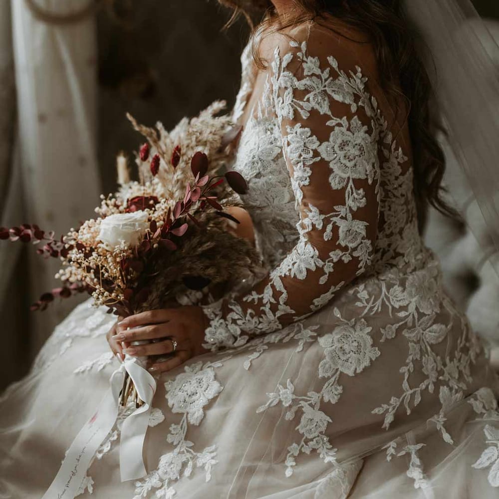 Bride wearing long sleeve lace illusion sleeve wedding dress with overlay holding boho dried flower bouquet 