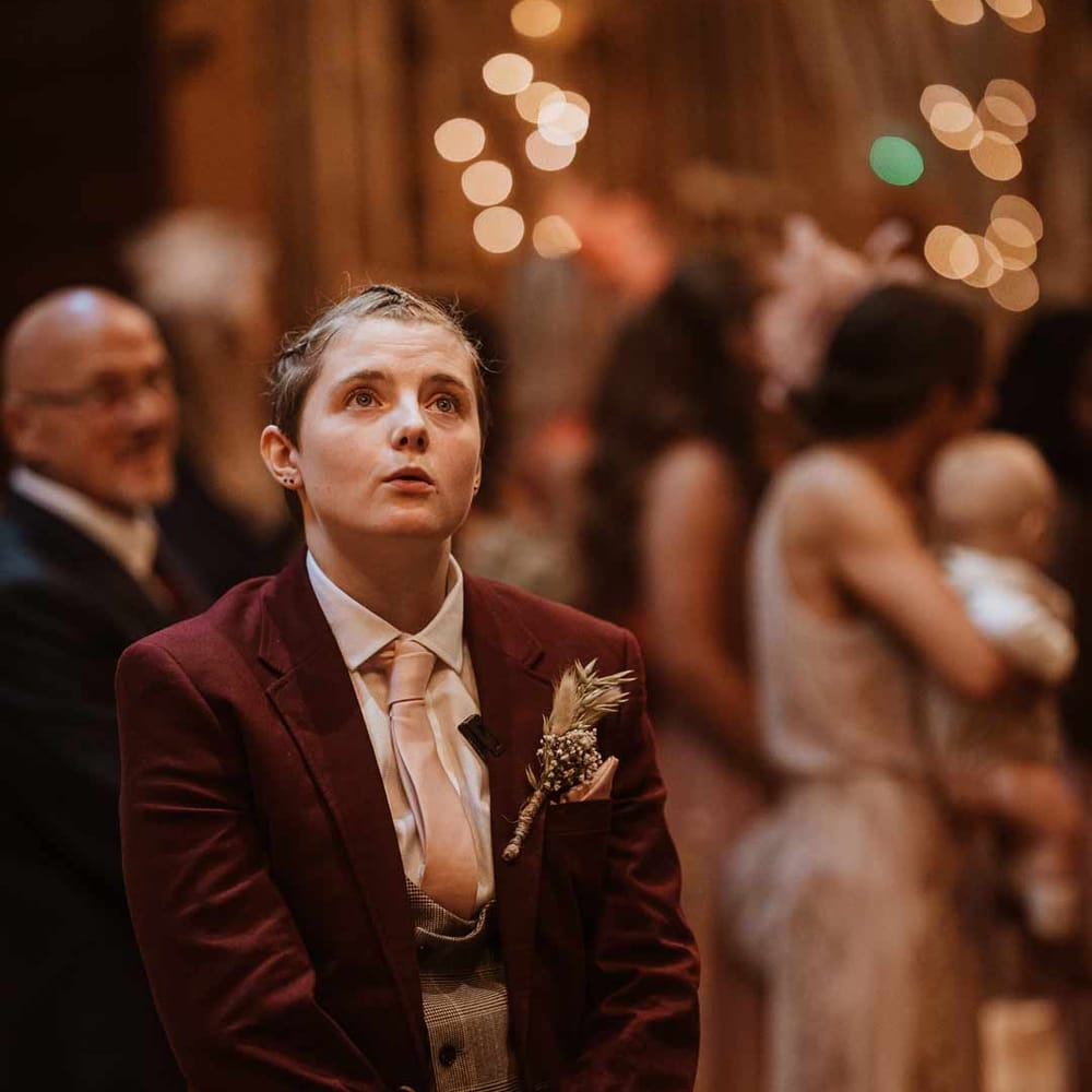 Bride in maroon bridal suit with grey waistcoat, dusky pink tie and autumnal dried flower boutonniere waiting at the end of the alter 