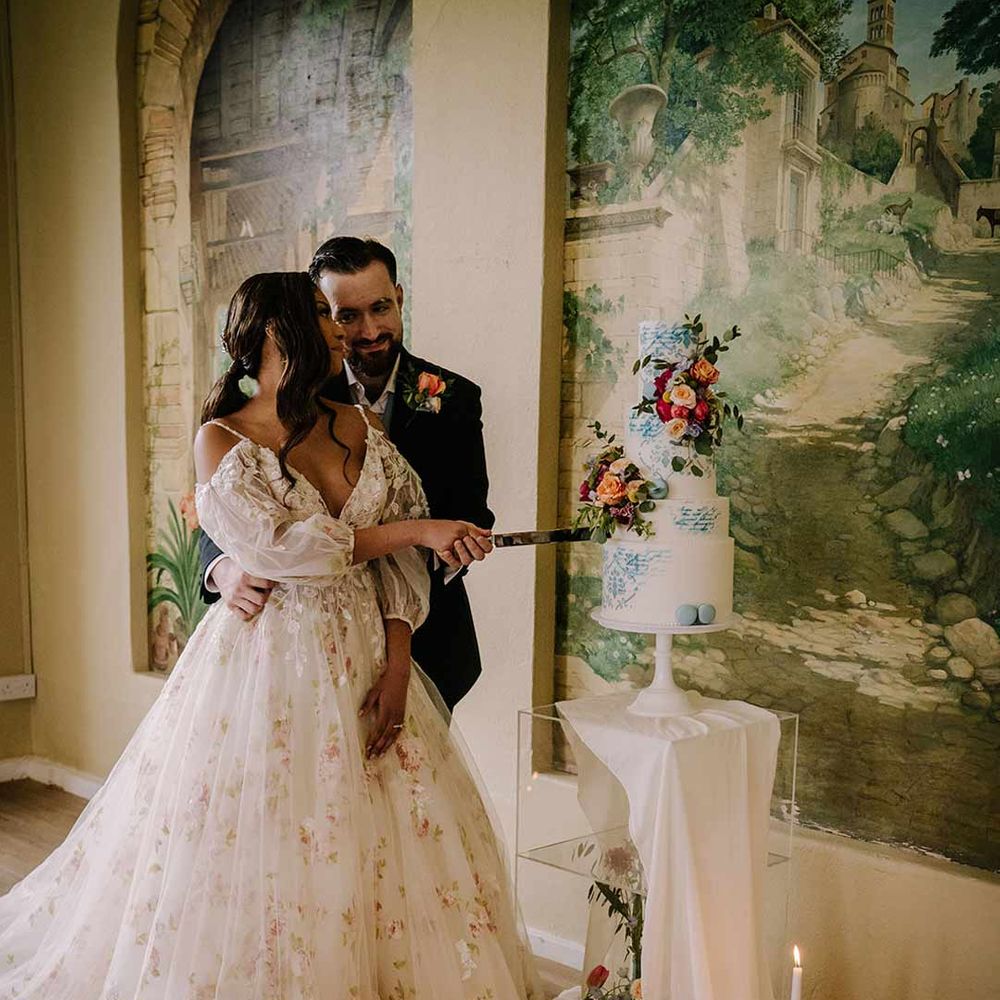 Groom wearing black blazer with pink garden rose and foliage boutonniere, cornflower blue waistcoat and black suit trousers cutting the wedding cake with bride in in v-neck long sleeve shoulder cut out 3d appliqué wedding dress with flecks of gold 