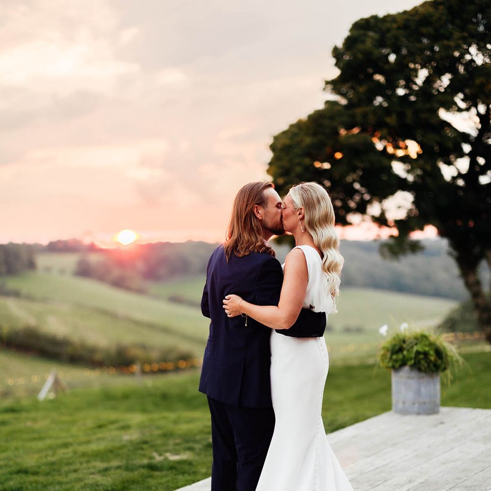 The bride in a Savin London wedding dress and groom in blue suit share a kiss during the sunset over the Surrey Hills 