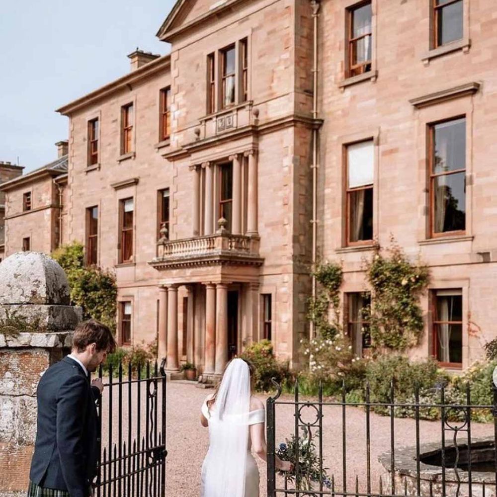 Bride in off shoulder satin wedding dress with church length bridal veil walking through the gate with groom in wedding kilt at Cambo House wedding venue Scotland 