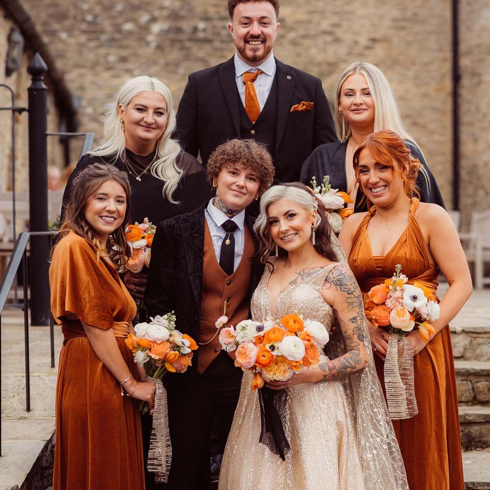 Bride in sleeveless deep v neck sparkly gold wedding dress with sheer and gold polka dot wedding veil holding orange garden rose, white poppy, light pink carnation and foliage bridal bouquet wrapped in sparkly gold ribbon standing with bride in dark velvet bridal jacket with rust waistcoat and pocket square, black suit trousers and gold pocket watch and bridesmaids in rust and black mismatched bridesmaid dresses and groomsmen in black suit with rust orange tie and pocket square at Kingscote Barn Tetbury 
