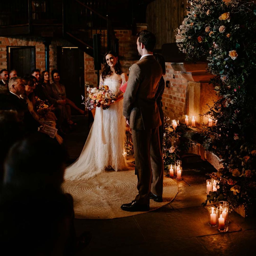 Bride in strapless ivory fringed crepe gown and chapel length veil holding mixed bridal bouquet with garden roses, carnations, peonies and foliage at the alter with groom in chocolate brown grooms suit at Willow Marsh Farm Loughborough 