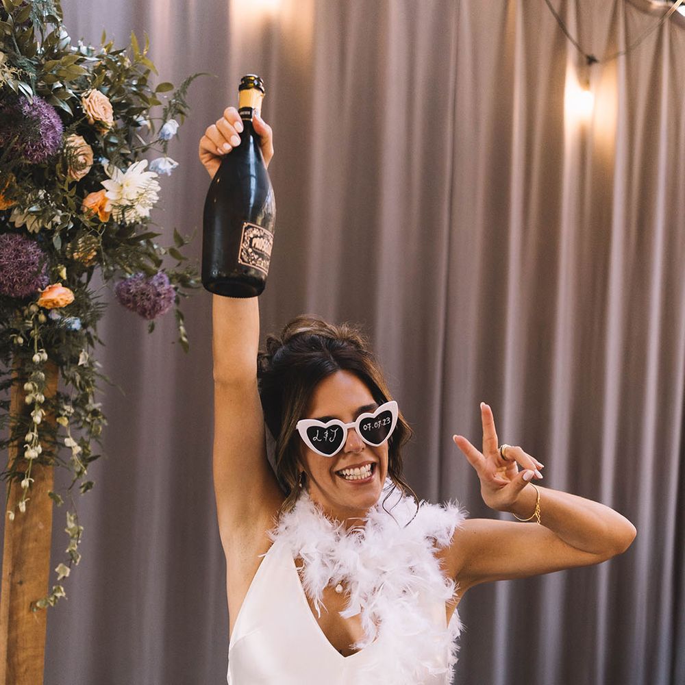 Bride in satin slip wedding dress with white feather boa wearing retro sunglasses and champagne bottle 