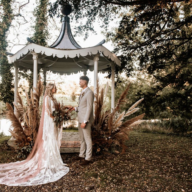 The bride and groom stand between pampas grass arrangements in front of the band stand