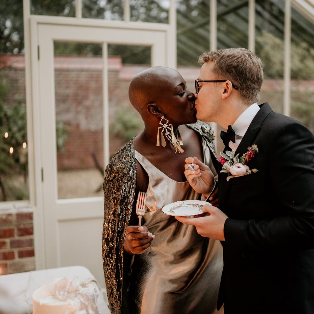 Groom in a tuxedo kissing his bride with alopecia in a black shimmering bridal cape 