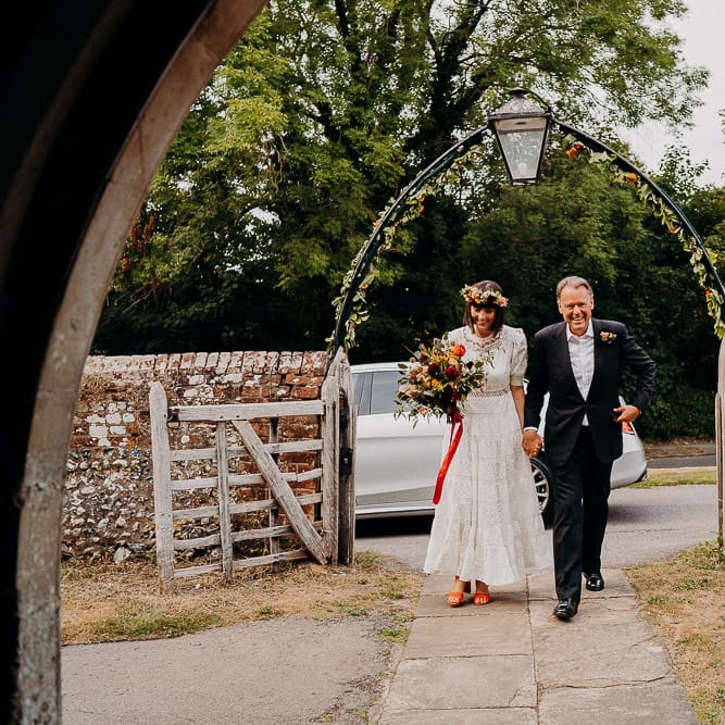 Bride in a white Broderie Anglaise wedding dress with colourful embroidery pattern arriving at the church with her father 