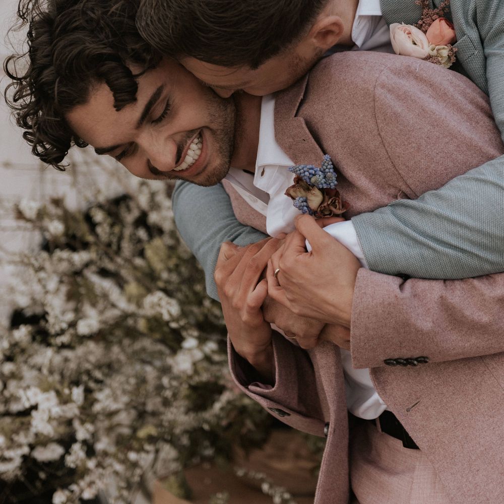 Grooms embrace whilst whilst wearing floral buttonholes 