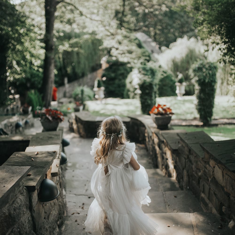 A flower girl in a pleated long white dress runs down steps.