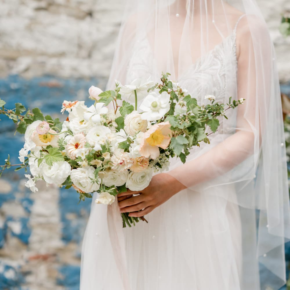 Beautiful bride in a delicate veil with pearl detail holding a white and yellow wedding bouquet with anemones, ranunculus and roses 