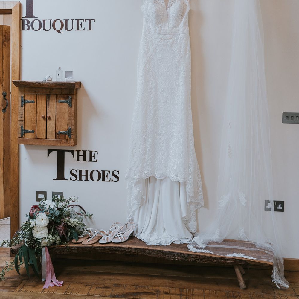 Lace wedding dress with train hung next to veil with lace trim, white heeled sandals, mixed bridal bouquet and sandals before Tythe Barn wedding with barn wedding flowers