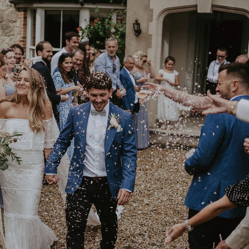 Bride in Grace Loves Lace bardot wedding dress and veil holding white and green bridal bouquet with macrame tie holds hands with groom in blue suit jacket and bow tie and guests throw confetti at Isle of Wight wedding with macrame wedding decor