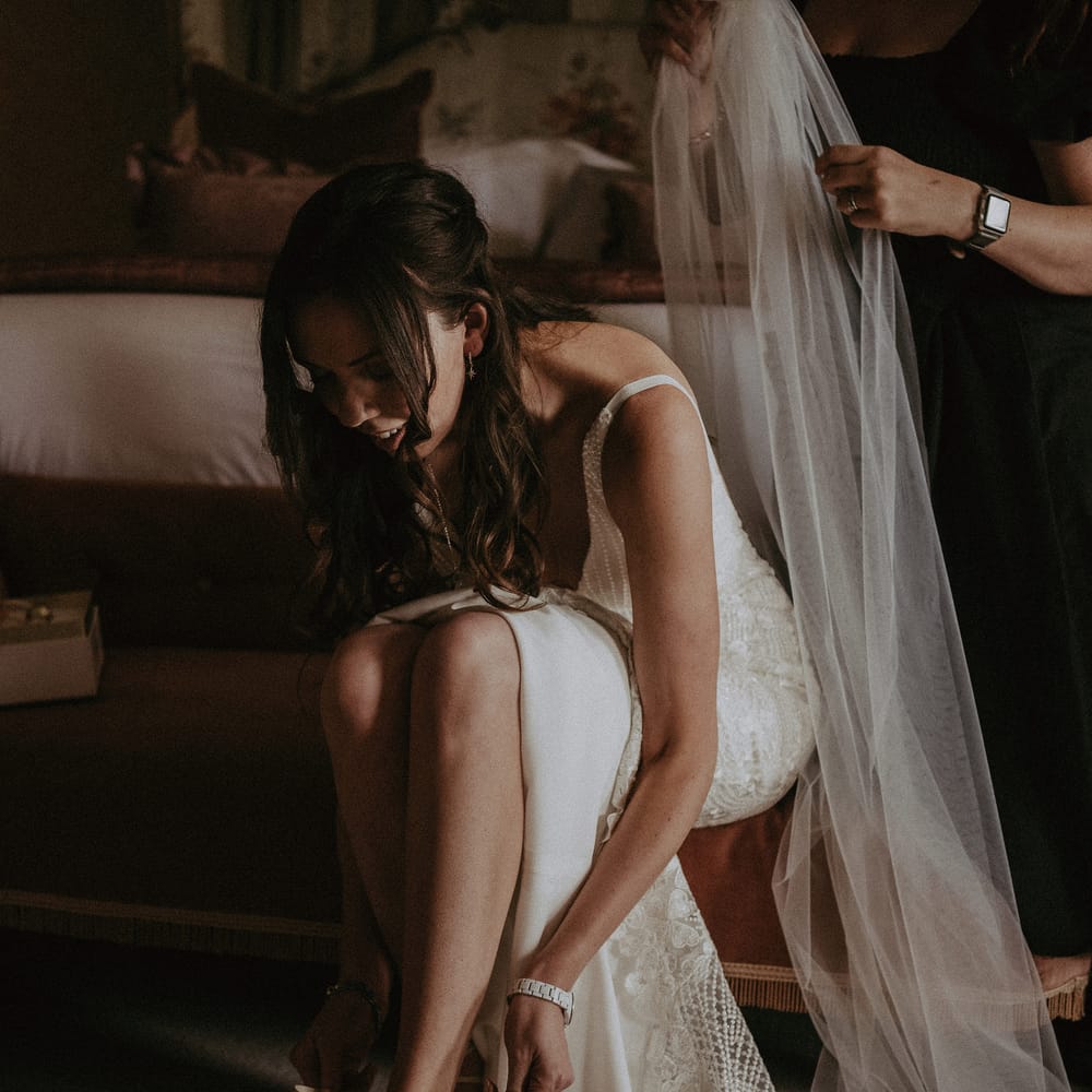 Bride ties her neutral shoes as her veil is slipped into her hair