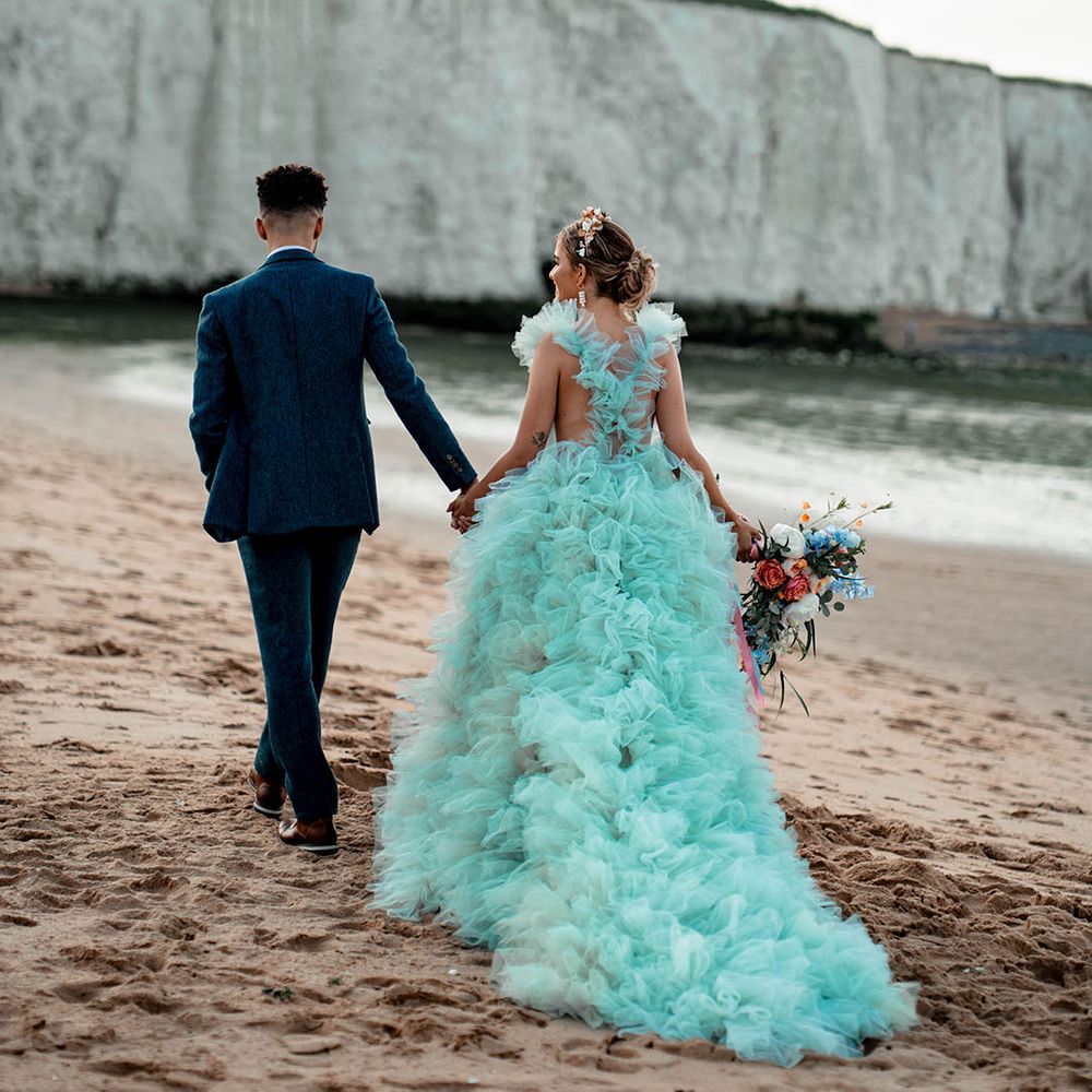 Bride in a Millia London mint green wedding dress walking along the beach with her husband in a blue wool suit 