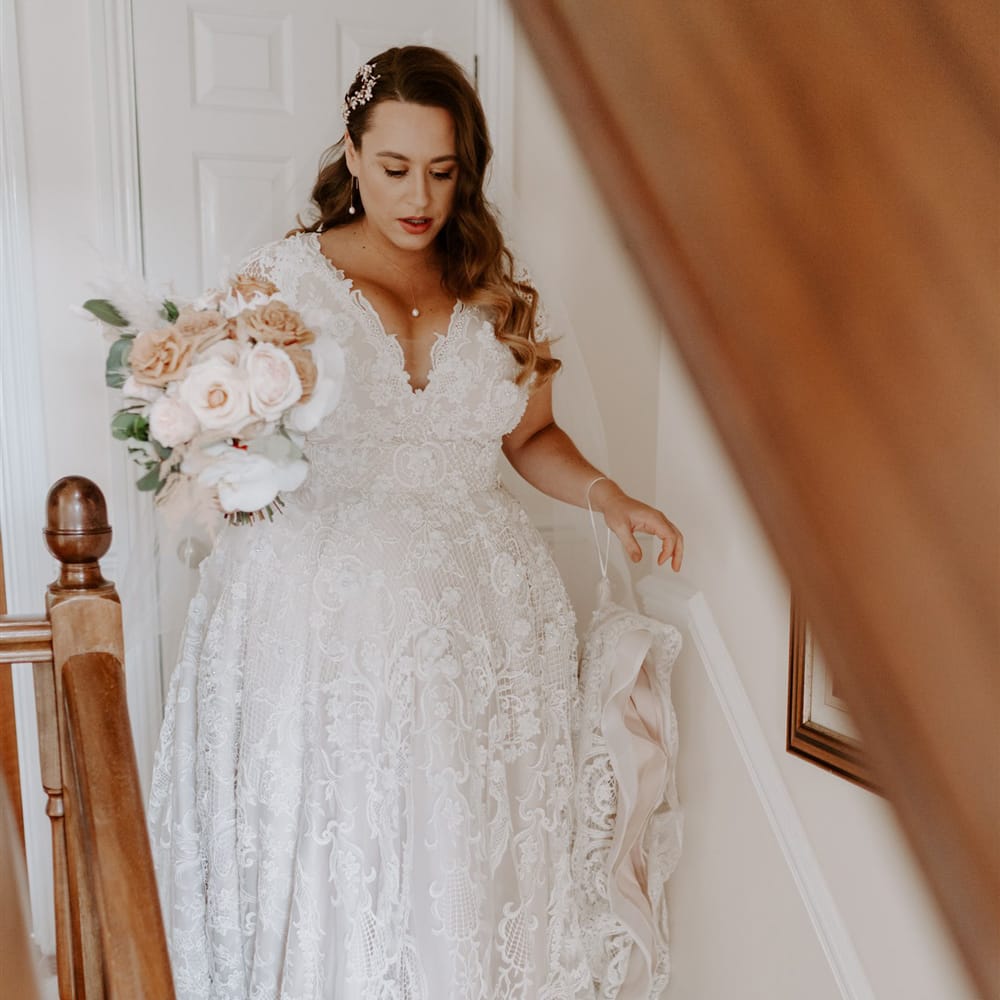 Bride in handmade wedding dress with mixed pink rose bridal bouquet walks down the stairs before DIY garden wedding in Bedfordshire