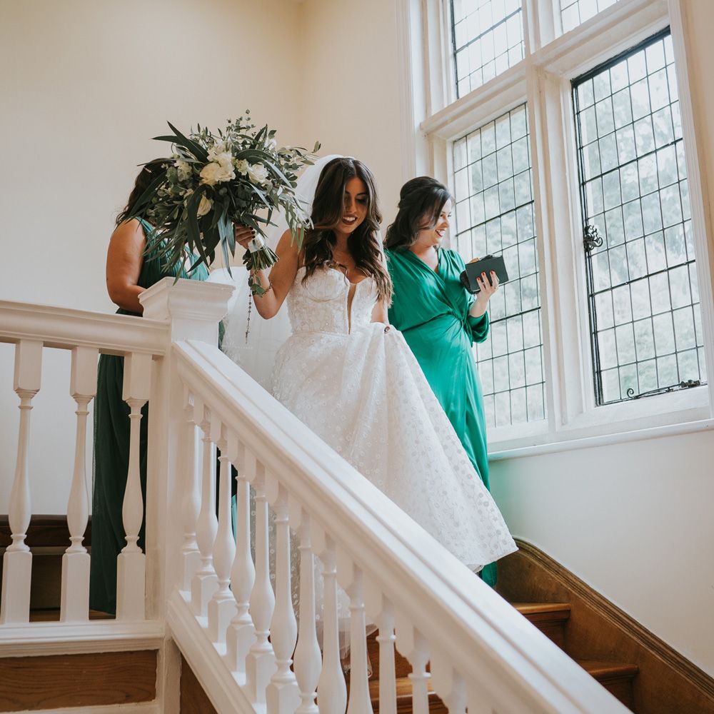 Bride in applique Pronovias wedding dress and Jimmy Choo wedding shoes walks down stairs holding green and white rose wedding bouquet with bridesmaids in green bridesmaid dresses at Notley Abbey before her wedding ceremony