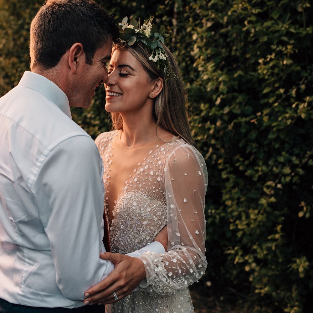 Bride in long mesh sleeve wedding dress with v neck and flower crown smiles whilst holding groom in white shirt and red tie during golden hour