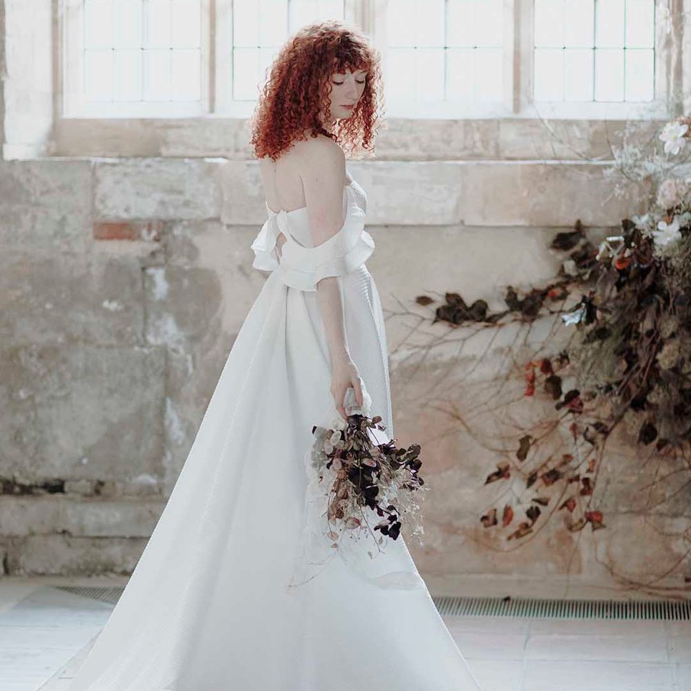 Bride with naturally curly red hair standing in Blackfriars Priory in a cold shoulder wedding dress holding a dried flower bouquet 