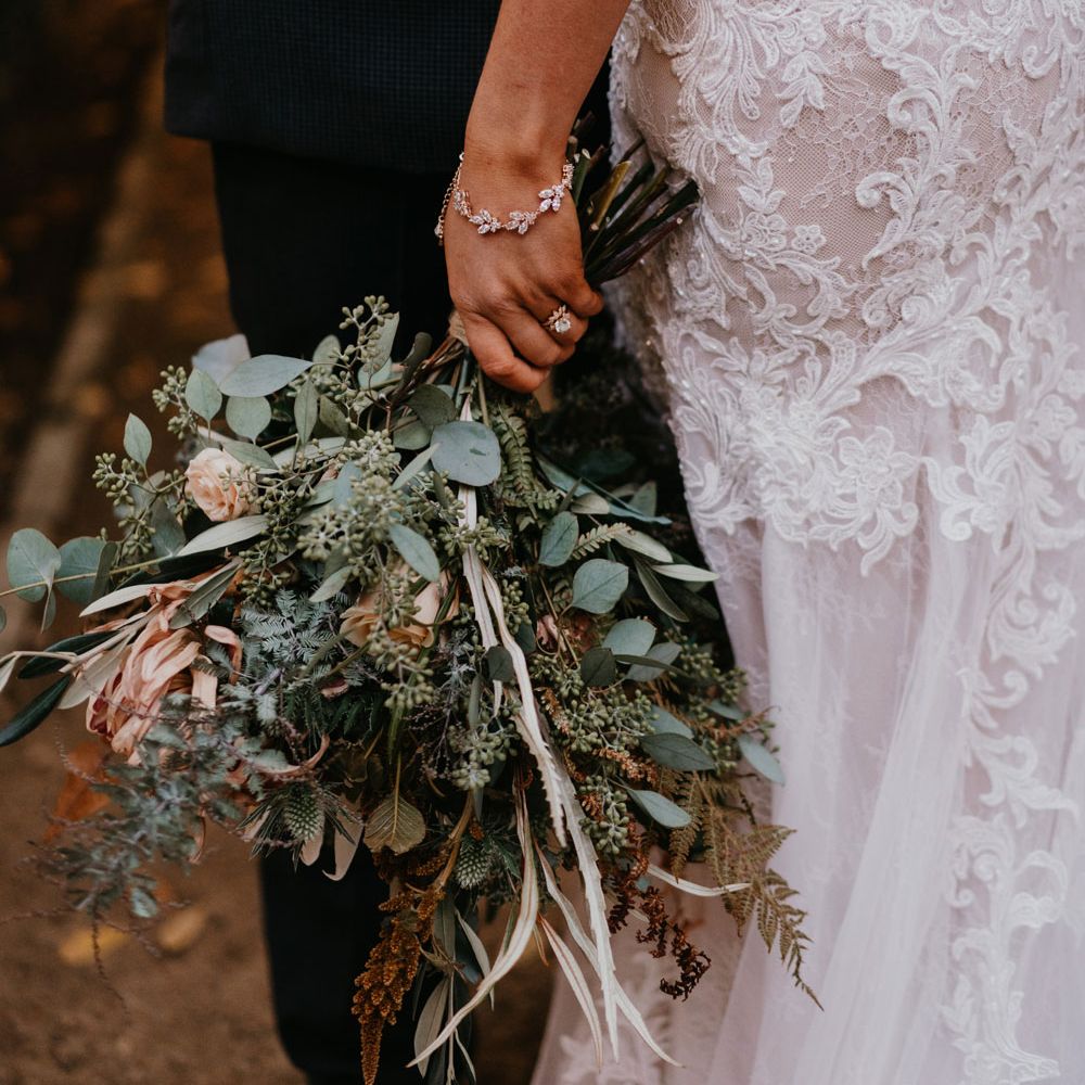 Bride in lace and tulle wedding dress wearing silver bracelet and large mixed bridal bouquet stands with groom in dark suit 