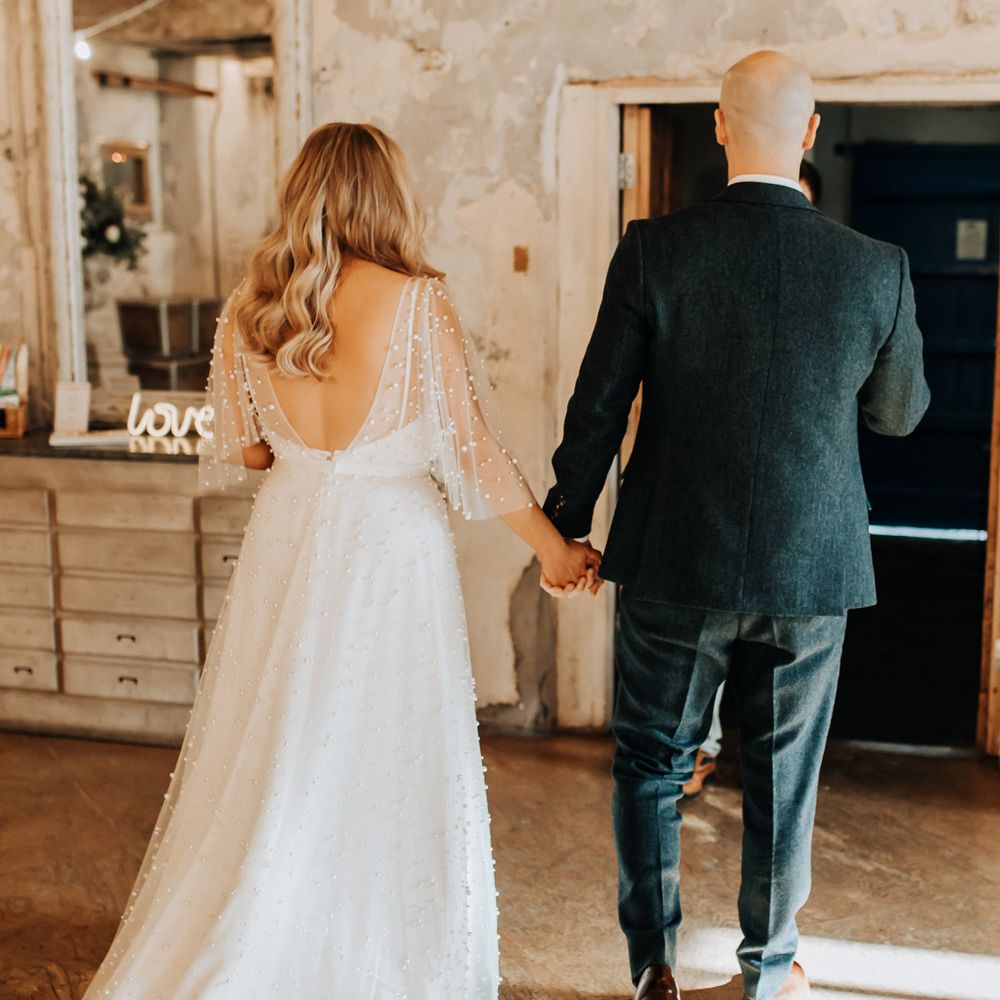 Bride in love v back wedding dress with pearls and train walks out of ceremony holding hands with groom in tweed suit and brown shoes
