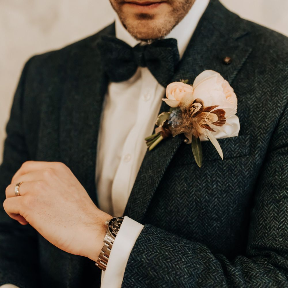 Groom in navy tweed three piece suit with bow tie and rose buttonhole adjusts his cuffs