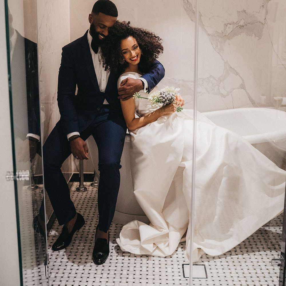 Black groom in a navy blue suit and bow tie embracing his Black bride in a one shoulder wedding dress in the bathroom at Mayfair Townhouse 