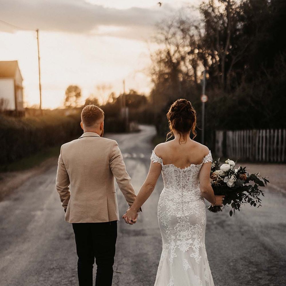 Bride in off the shoulder lace wedding dress with button details and mermaid style with groom in beige suit jacket 