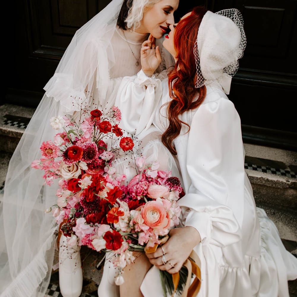 Bride with red wavy hair in a long sleeve wedding dress kissing her bride in a high neck wedding dress with long veil 