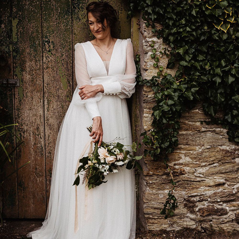 Bride leans against a wall wearing sheer long sleeve wedding dress by Sassi Holford