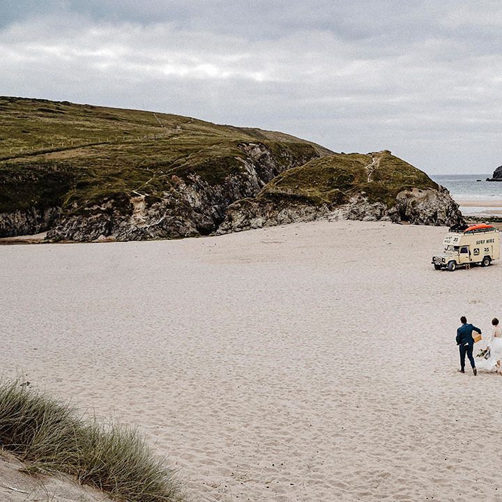 Bride and groom walk along Cornwall beach together with van in distance