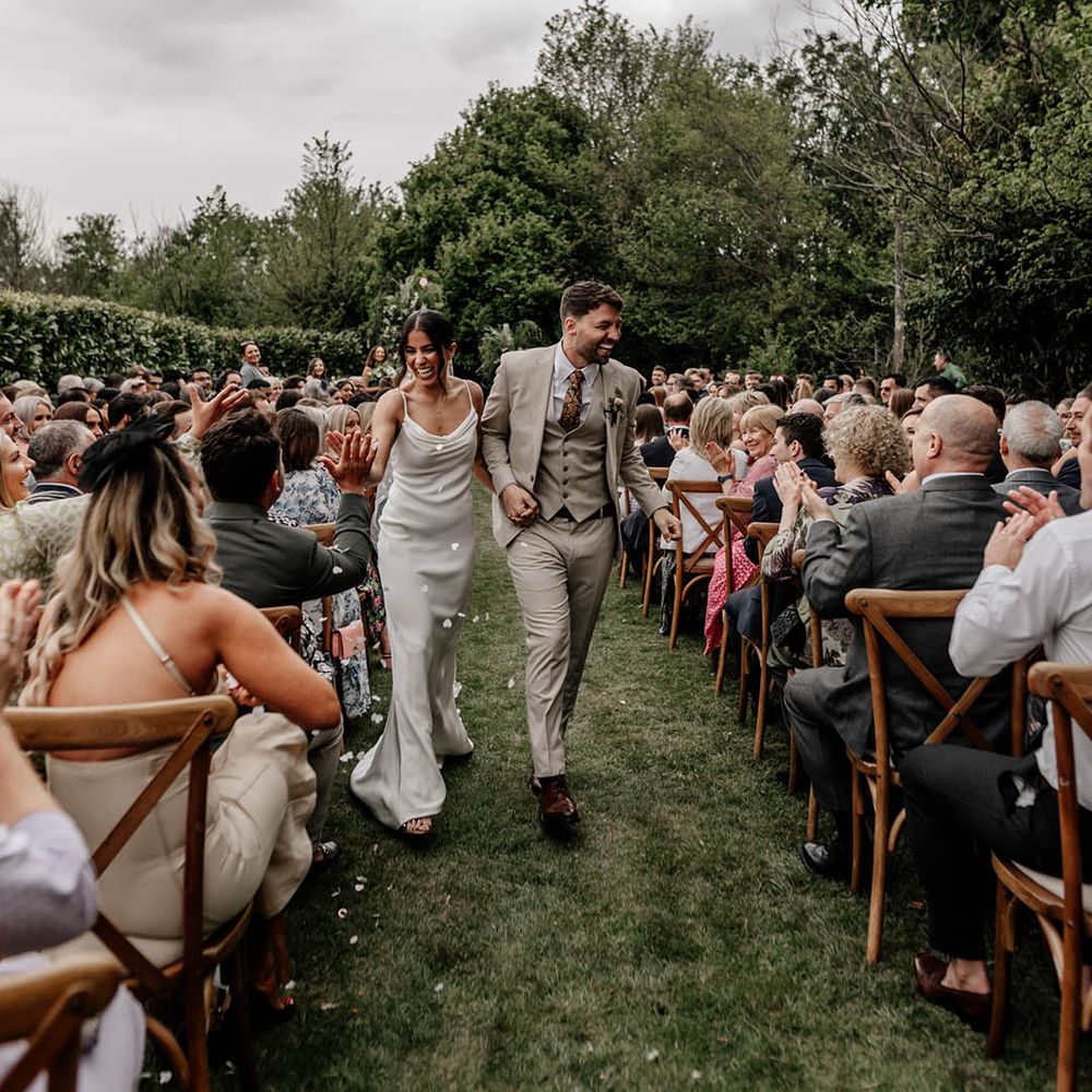 Groom in a stone coloured suit holding descending up the aisle with his bride in a satin slip wedding dress and pearl veil at their outdoor back garden wedding