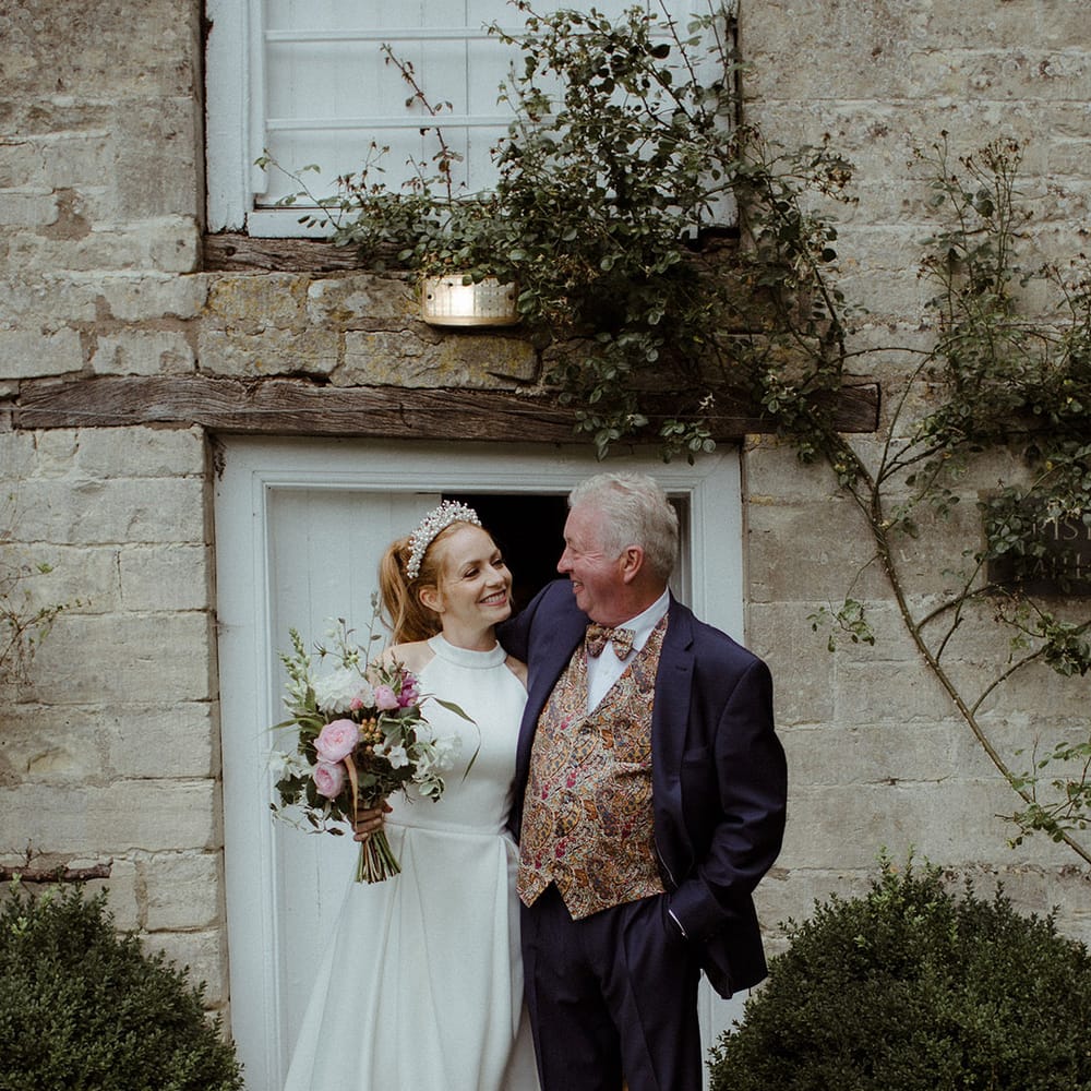 Bride in pleated wedding dress with pearl headband stands with groom in paisley patterned waistcoat and tie 