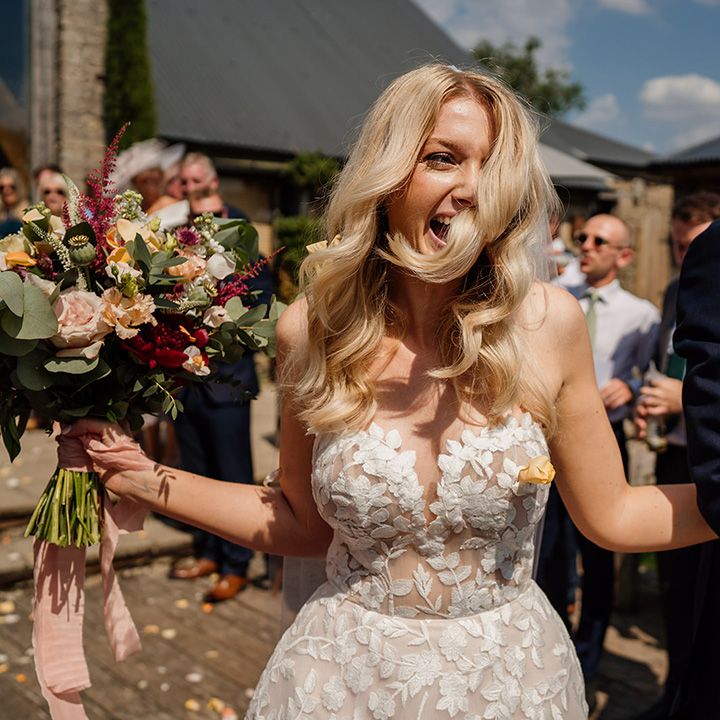 Bride with softly curled blonde hair in strapless lace Lillian West wedding dress walks through confetti with groom in blue suit 