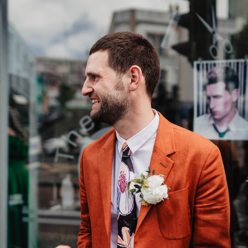 Groom wears orange Paul Smith linen suit complete with white shirt, floral illustrated tie and white floral buttonhole 
