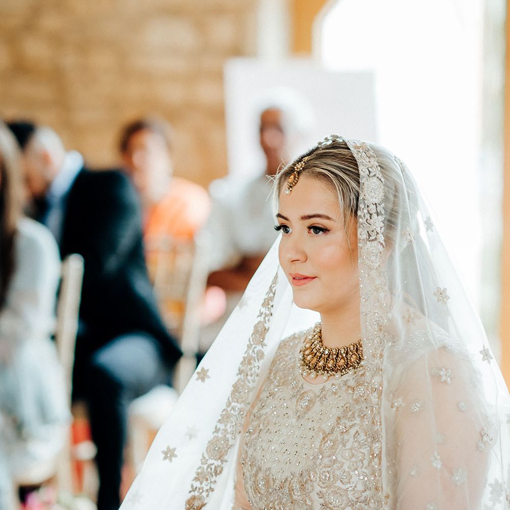 Bride wears her blonde hair tied back beneath embellished sheer veil complete with gold necklace and pearl drops 
