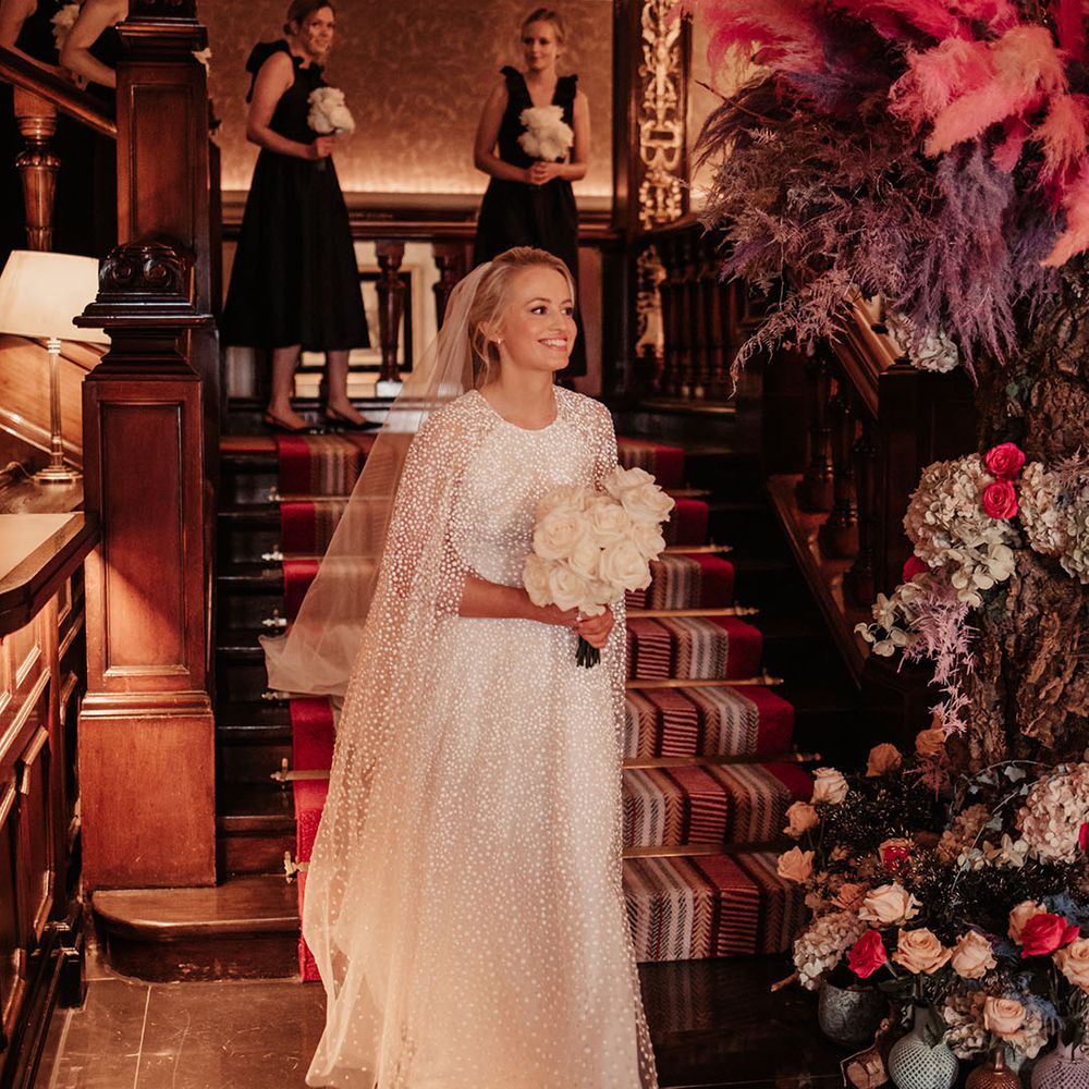 Bride in sparkly Jesus Peiro cape and wedding dress with white rose buttonhole walking next to colourful pampas grass decor 