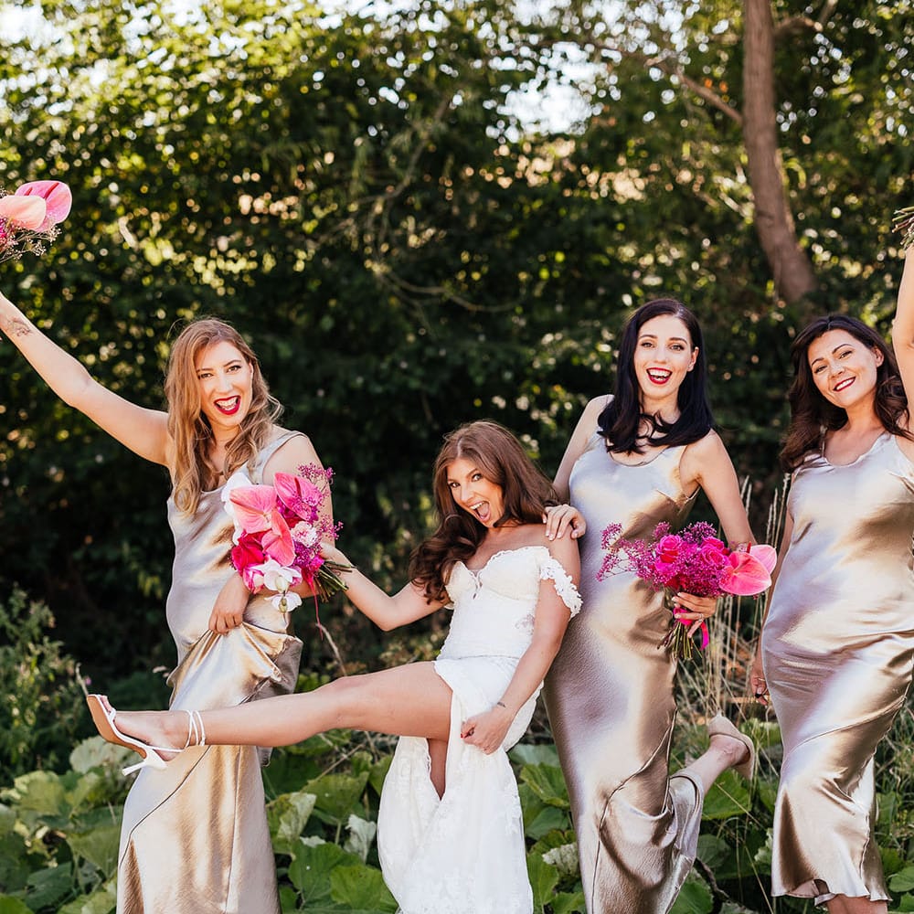 Bride stands with her bridesmaids who wear champagne bridesmaid dresses whilst holding bright floral bouquets 
