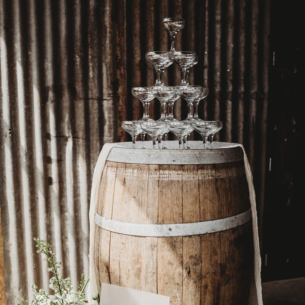Champagne tower wedding decor on a wooden barrel with a frosted acrylic sign, drapery and white flowers 