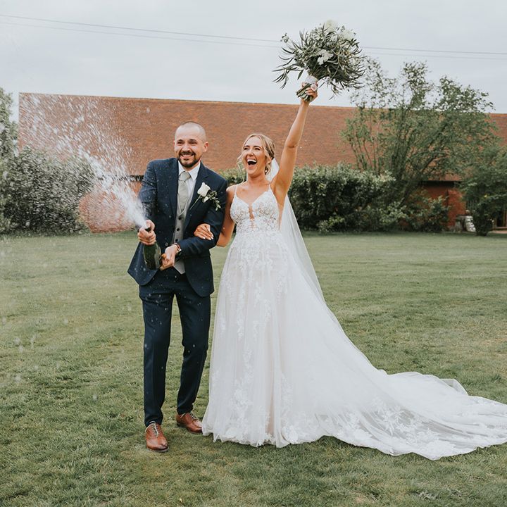 Groom in dark blue wedding suit pops and sprays a bottle of champagne with the bride 