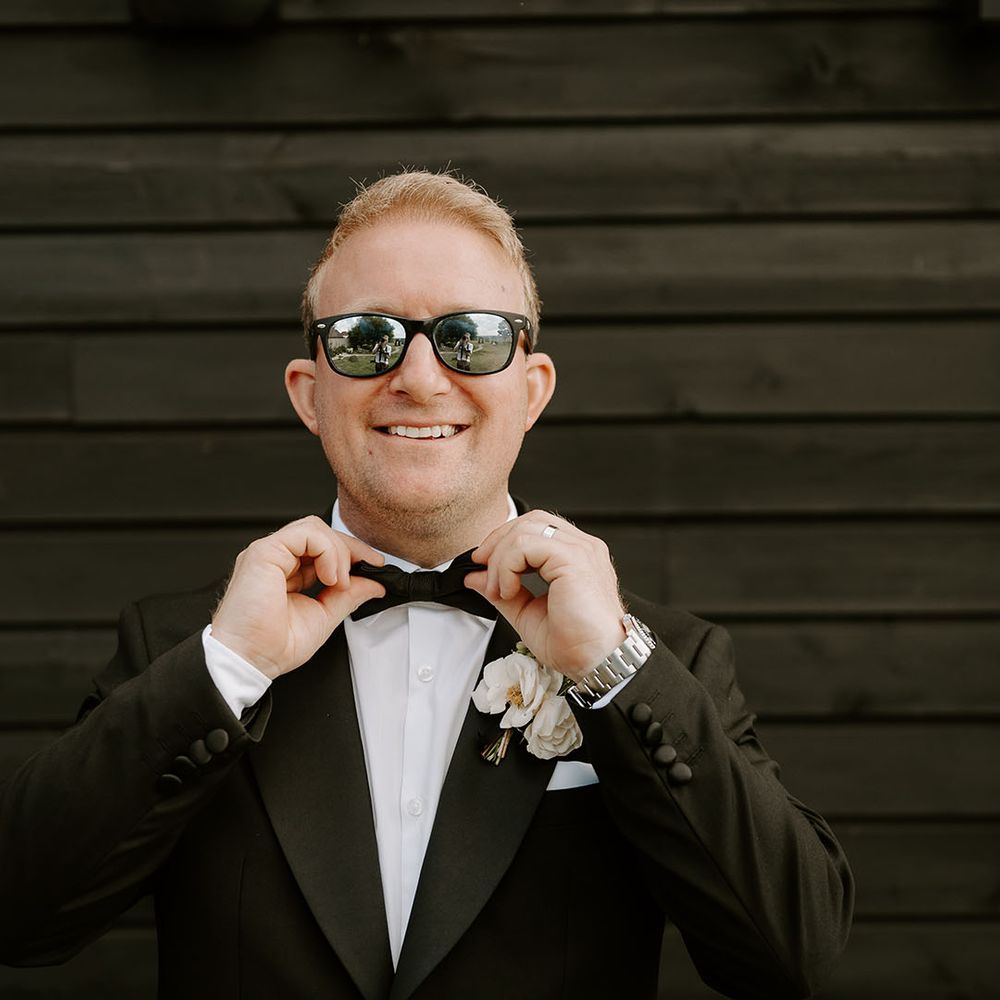 Groom wears black tie with sunglasses and white floral buttonhole 